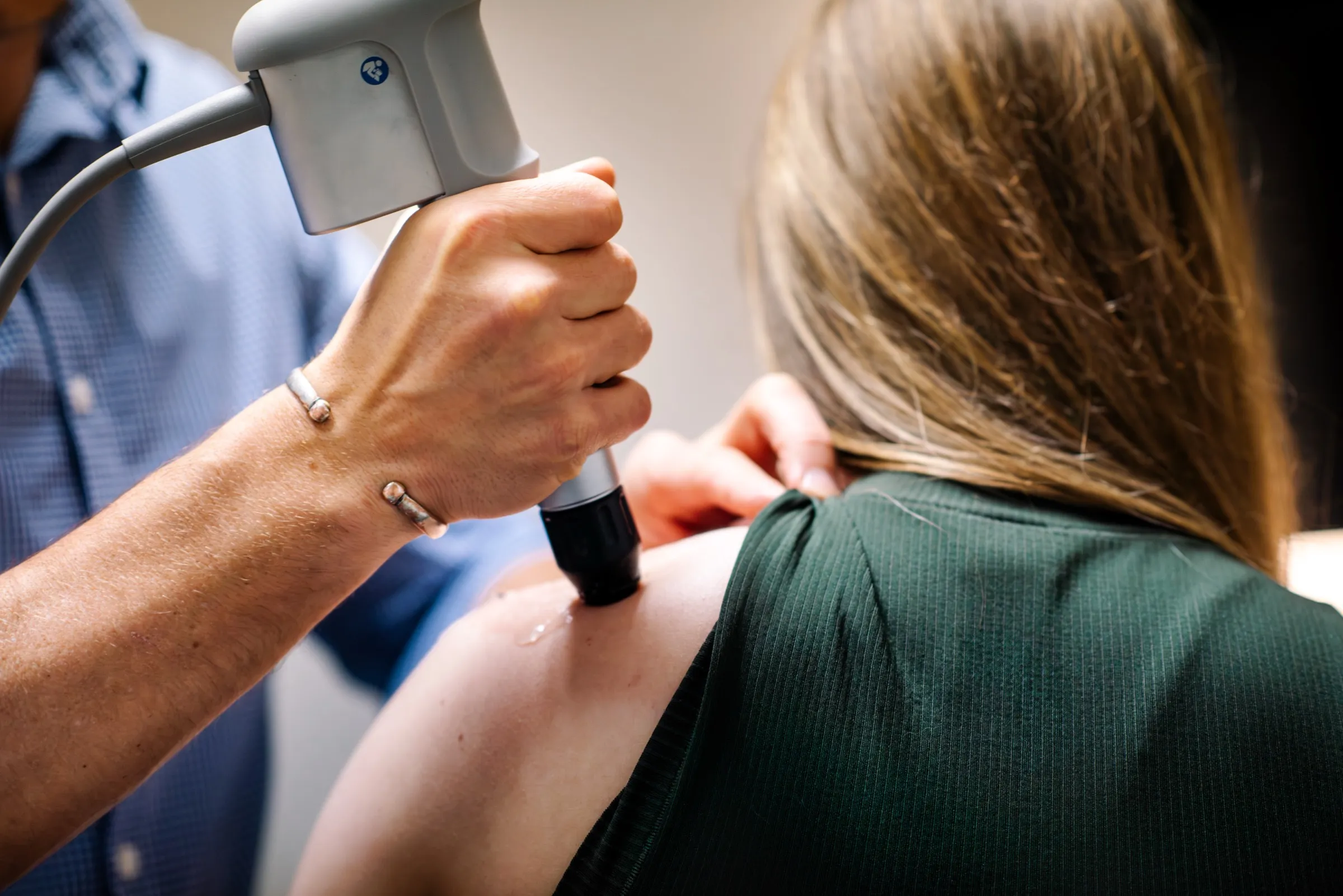 A clinician applying the shockwave handpiece, with gel, to the back of a seated patient's shoulder near the shoulder blade
